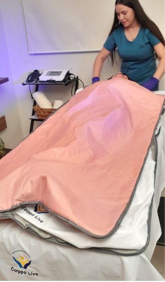 Una paciente se somete a un tratamiento de envoltura corporal en la clínica Corppo Live. Una profesional de la salud, vestida con uniforme azul y guantes, ajusta la manta térmica rosada que cubre a la paciente, quien está recostada sobre una camilla con una sábana blanca. En el fondo, se puede ver un equipo médico y toallas enrolladas sobre una mesa auxiliar.